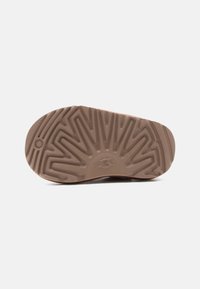 Καφέ σόλα παπουτσιού UGG με εγκοπές σε ζιγκ ζαγκ και κυκλικό σχέδιο κοντά στη φτέρνα σε απλό λευκό φόντο.