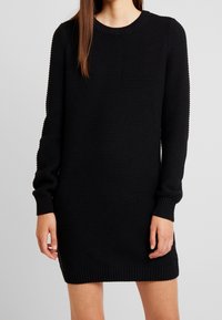 Abito nero lavorato a maglia con maniche lunghe, scollatura rotonda, e polsini e orlo a costine. Tessuto testurizzato con vestibilità ampia.