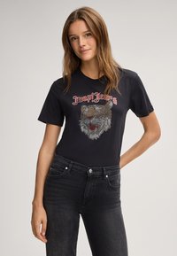 T-shirt noir à manches courtes présentant un graphisme de tête de tigre en tons argent et rouge, avec le texte « Dope! Jeans » au-dessus du design.