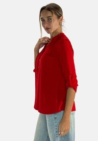 Camiseta roja de ajuste holgado, con botones en la parte frontal, bolsillo en el pecho y mangas enrollables, combinada con jeans azul claro de tela texturizada.