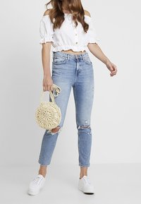 Blusa corta blanca con detalles de botones, jeans rasgados azul claro y bolso redondo de mimbre en color crema. Modelo lleva zapatillas blancas.