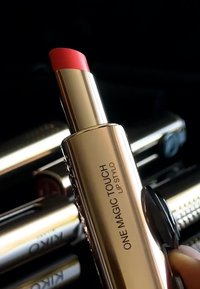Kultaputkinen huulipuna, jossa on vaaleanpunainen-punakotelo, jossa on kierrettävä mekanismi ja kaiverrettu etiketti "ONE MAGIC TOUCH LIP STYLO." Silkkinen koostumus.