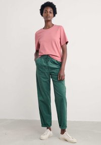 Różowa bawełniana koszulka z krótkimi rękawami w połączeniu z zielonymi spodniami na sznurek. Całość uzupełniają kremowe buty casualowe.