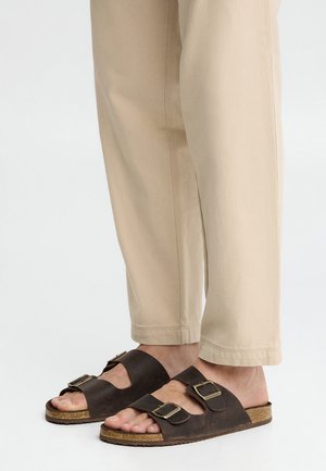 Füße in beigen Hosen und dunkelbraunen Ledersandalen mit doppelter Schnalle und Korksohlen, stehend auf einer weißen Fläche.