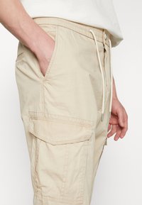 Pantaloni cargo beige realizzati in tessuto leggero, dotati di vita con cordino, tasche laterali e una finitura textured.