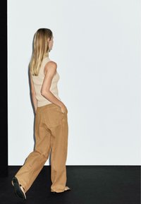 Woman with long blonde hair walking away, wearing beige sleeveless top and loose brown pants with hands in pockets on black and white floor.