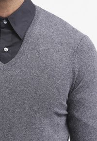 Szary sweter z dzianiny z dekoltem w serek, charakteryzujący się miękką fakturą i prążkowanymi zdobieniami na dole i mankietach, noszony na czarnej koszuli.