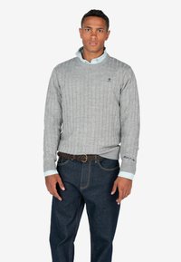 Pullover grigio a maglia con una camicia blu con colletto sotto, abbinato a jeans blu scuro e una cintura intrecciata marrone.