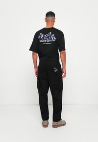 Man die met zijn rug naar de kijker staat, gekleed in zwarte cargobroek, zwart graphic t-shirt met de tekst "Alpha Studio Edition" en beige laarzen.
