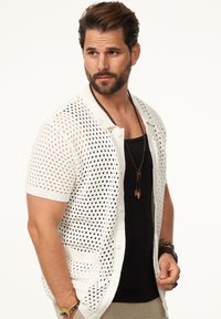 Weißer, offen gestrickter Cardigan mit kurzen Ärmeln, ausgestattet mit einer Knopfleiste und zwei Taschen. Über einem schwarzen Tanktop getragen, das ein strukturiertes Gewebe zeigt.