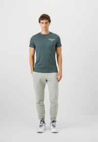 Άνδρας στέκεται μπροστά σε απλό φόντο φορώντας ένα σκούρο πράσινο μπλουζάκι Björn Borg, ανοιχτού γκρι παντελόνι φόρμας και λευκά αθλητικά παπούτσια με ναυτικές λεπτομέρειες.