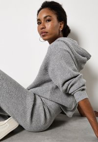 Szary hoodie o fakturze wafelka z kapturem i podwiniętymi rękawami, zestawiony z pasującymi szarymi spodniami dresowymi i białymi sneakersami, ustawiony przy ścianie.