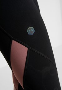 Černé sportovní legíny s růžovým panelem, vyznačující se hexagonálním logem a kontrastním šitím. Hladká textura, přiléhavý design.