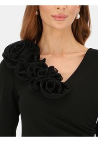 Czarna sukienka z dekoltem w serek i dużym, teksturowanym falbanem na lewym ramieniu, dodającym wymiaru i detali.