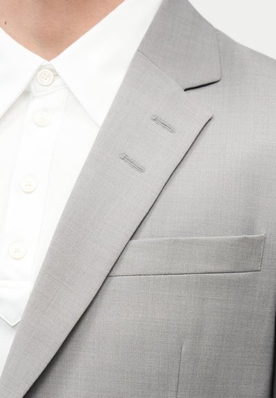 Κοντινή λήψη του πέτου σακακιού ανοιχτόγκρι κοστουμιού και της τσέπης στο στήθος πάνω από λευκό πουκάμισο με γιακαδάκι κλεισμένο με κουμπί.