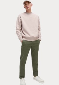 Helles rosa Sweatshirt mit Rundhalsausschnitt und gerippten Bündchen, kombiniert mit grünen, schmal zulaufenden Hosen und weißen Sneakers. Einfaches Design, lässiger Stil.