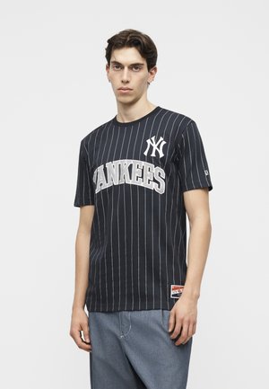 Νέος άνδρας που φοράει μαύρο ριγέ μπλουζάκι των Yankees και μπλε τζιν, στέκεται με ουδέτερη έκφραση μπροστά σε απλό φόντο.