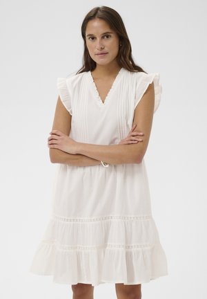 Žena s dugom smeđom kosom koja nosi bijelu haljinu bez rukava s naborima i čipkom, prekriženih ruku, neutralnog izraza, stojeći na bijeloj pozadini.