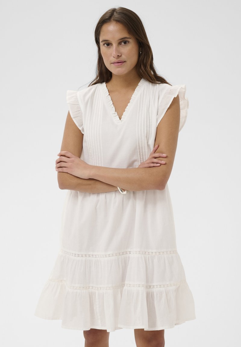 Femme aux longs cheveux bruns portant une robe blanche sans manches avec des volants et de la dentelle, les bras croisés, expression neutre, debout sur un fond blanc.