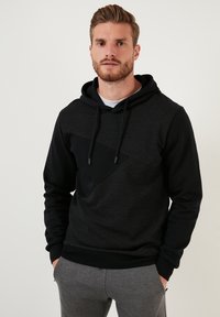 Sudadera con capucha negra con un panel diagonal texturizado, que presenta cordones y puños acanalados. Llevada con pantalones deportivos grises.