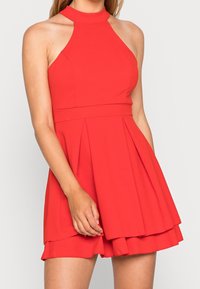Czerwona sukienka bez rękawów z wysokim dekoltem, dopasowanym korpusem i rozkloszowanym, plisowanym dołem. Wykonana z gładkiego materiału z warstwowym wykończeniem. 