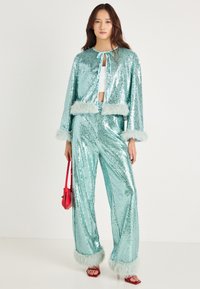 Conjunto de duas peças em turquesa com lantejoulas, composto por uma jaqueta cropped e calças de perna larga, com detalhes em pele sintética nas mangas e na bainha. Acessório de bolsa vermelha.