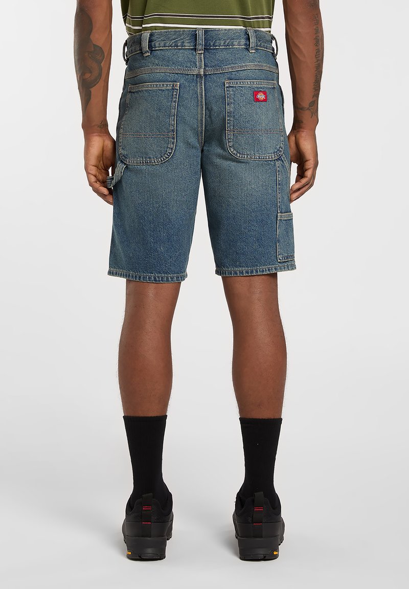 Uomo che indossa pantaloncini da lavoro in denim blu con grandi tasche posteriori e anello per martello, abbinati a calzini e scarpe nere, visto da dietro.