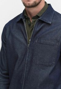 Man som bär en mörkblå zip-up-jacka över en olivgrön skjorta med krage, vilket visar bröst- och halsområdet.