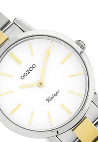 Montre argentée et dorée avec un cadran blanc, comportant des marqueurs et des aiguilles dorés, ainsi que le texte "OOZOO Vintage". Le bracelet métallique a des accents dorés.