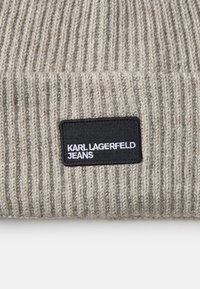 Bež ribaga kudum, mille esiosas on must ristkülikuline silt valge tekstiga "KARL LAGERFELD JEANS".