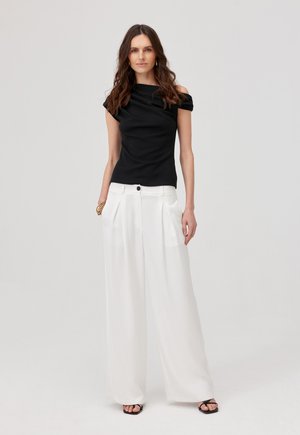 Kobieta z długimi, brązowymi włosami nosi czarny top odsłaniający ramiona, szerokie białe spodnie z wysokim stanem, czarne sandały i złote bransoletki.