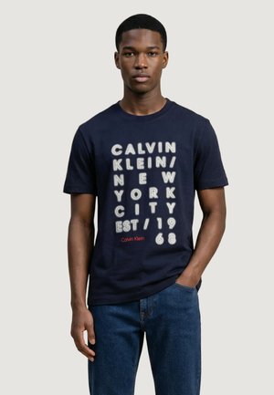 Młody mężczyzna ubrany w granatowy t-shirt marki Calvin Klein z białym napisem "Calvin Klein/New York City/Est. 1968" oraz niebieskie dżinsy.