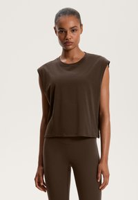 Barna, rövidített ujjatlan felső, puha pamutból készült, kerek nyakkivágással és laza szabással. Hozzá illő barna leggings-szel párosítva.