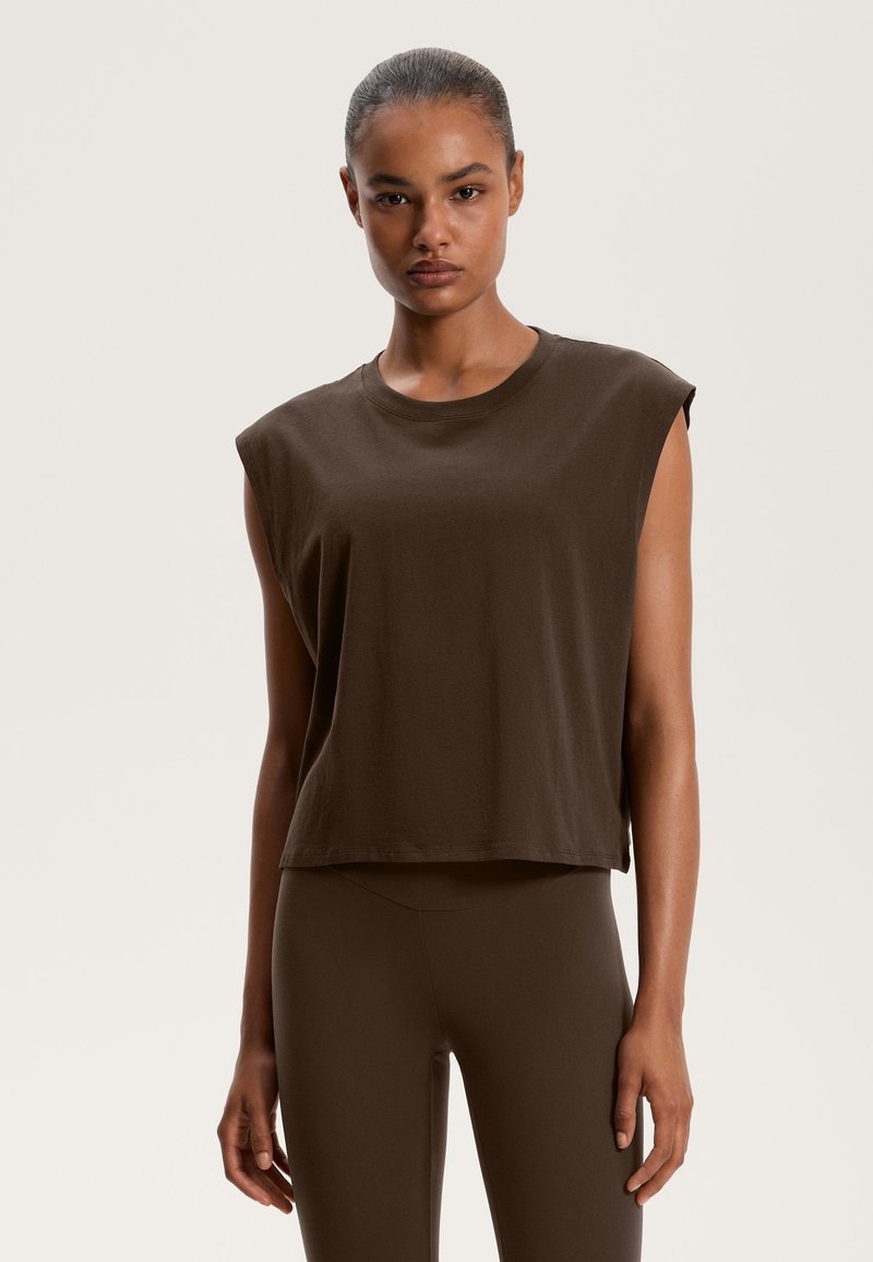 Barna, rövidített ujjatlan felső, puha pamutból készült, kerek nyakkivágással és laza szabással. Hozzá illő barna leggings-szel párosítva.