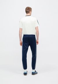 Fehér atlétikai póló fekete gallérral, kék melegítőnadrággal, oldalcsíkkal. Kék cipő teszi teljessé a sportos megjelenést.