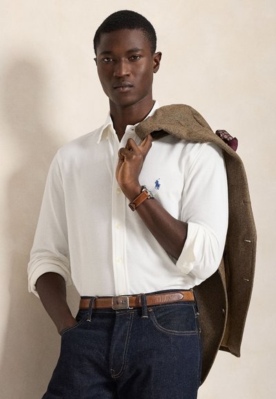 Jeune homme portant une chemise blanche boutonnée et un jean foncé, tenant une veste marron sur l'épaule, debout devant un fond beige uni.