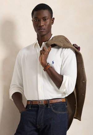 Jeune homme portant une chemise blanche boutonnée et un jean foncé, tenant une veste marron sur l'épaule, debout devant un fond beige uni.