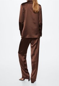 Hnědá saténová blůza s dlouhým rukávem a volným střihem, doplněná odpovídajícími širokými kalhotami a černými sandály na podpatku, viděná zezadu.