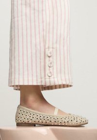 Ballerine in suede beige con embellishments di borchie argentate. Abbinate a pantaloni a righe rosa e bianche con dettagli di bottoni.