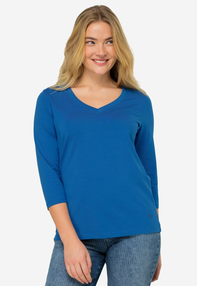 Modrá dlouhá vesta s výstřihem do V a hladkou texturou. V blízkosti lemu má nenápadné logo a je stylizována s upnutými džíny.