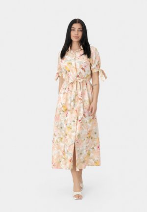 Donna con capelli lunghi e neri che indossa un vestito midi floreale pastello con bottoni, maniche annodate e cintura in vita, in piedi e rivolta in avanti.