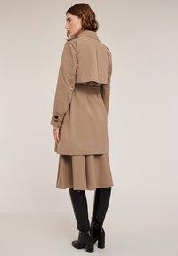 Světle hnědý trench coat s krátkým střihem, páskem v oblasti pasu a knoflíkovými rukávy. V kombinaci s volnou béžovou sukní a černými kozačkami nad kolena.