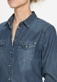 Camicia di denim blu scuro, con chiusura a bottoni frontalmente, due tasche sul petto e colletto classico. Texture morbida con cuciture visibili.