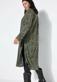Camisa larga verde oliva con estampado de leopardo negro, que presenta un corte holgado, mangas largas, aberturas laterales, y se lleva sobre leggings negros y botas con tachuelas.