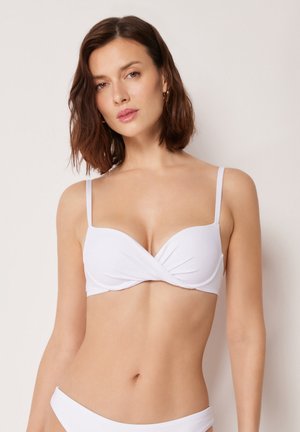 Kvinde med skulderlangt brunt hår iført hvid bikini top og matchende bund mod en ensfarvet lys baggrund.