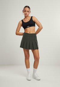 Schwarzes Cropped-Top und dunkelgrüne, plissierte Hose. Die Hose hat eine hohe Taille und seitliche Details. Das Model trägt weiße Sneaker und kurze Socken.