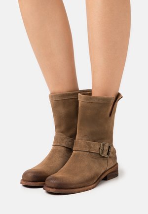 Bruine suède enkellaarsjes met zijriempjes en gespen, gedragen op blote benen, met afgeronde neuzen en lage houten hakken.