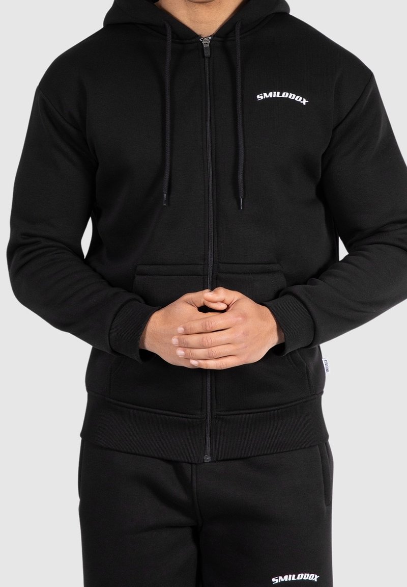 Sweatshirt à zip noir en mélange de coton, avec une poche avant, des cordons de serrage, et un logo blanc sur la poitrine. Tissu texturé et poignets ajustés.