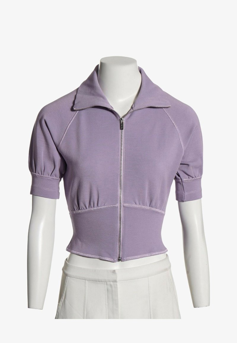 Haut court lavande à manches courtes avec fermeture éclair et coutures froncées, porté sur un buste de mannequin blanc avec un pantalon blanc.