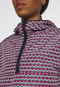 Leichte, gemusterte Jacke mit einem geometrischen Design in Rot, Weiß und Marineblau, mit Reißverschluss und Kapuze. Sichtbare Textur und Nähedetails.